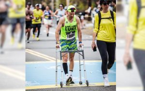 Man wearing green athletic-wear that reads "Live Like Luke" walking in marathon with help from a walker