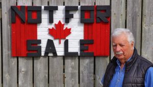 Man stands beside his wooden tobacco slat sign that reads "Not for sale" on a Canadian flag